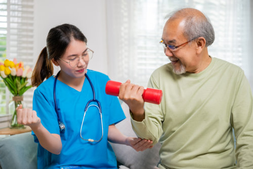 A caregiver in blue scrubs demonstrates an arm exercise while helping an elderly man who is smiling and lifting a red dumbbell