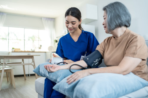 A caregiver in a blue uniform sits on a sofa, carefully checking the blood pressure of an elderly woman who is resting her arm on a blue pillow