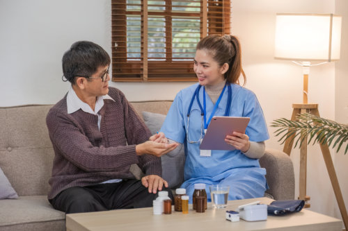 A healthcare worker in blue scrubs holding a tablet smiles while assisting an elderly man, who is sitting on a sofa and holding medication in his hand, with various pill bottles and a glass of water on the coffee table in front of them
