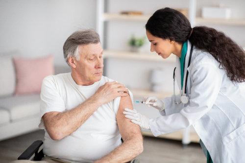 A doctor in a white coat and gloves administers an injection into the shoulder of an elderly man sitting in a chair