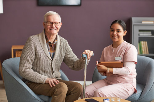 Elderly man with a cane smiling next to a seated female nurse holding a clipboard in a cozy room.