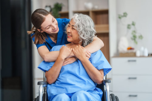 Young female caregiver embraces and smiles at elderly woman in wheelchair in a bright, homey indoor setting.