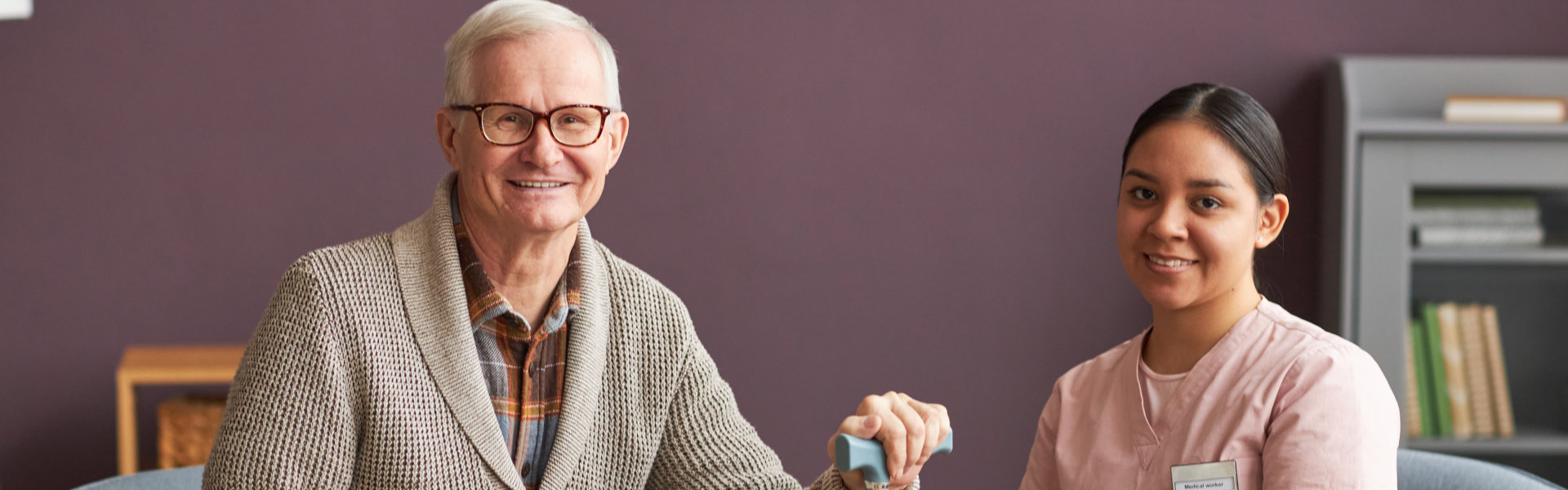 Elderly man with a cane smiling next to a seated female nurse holding a clipboard in a cozy room.