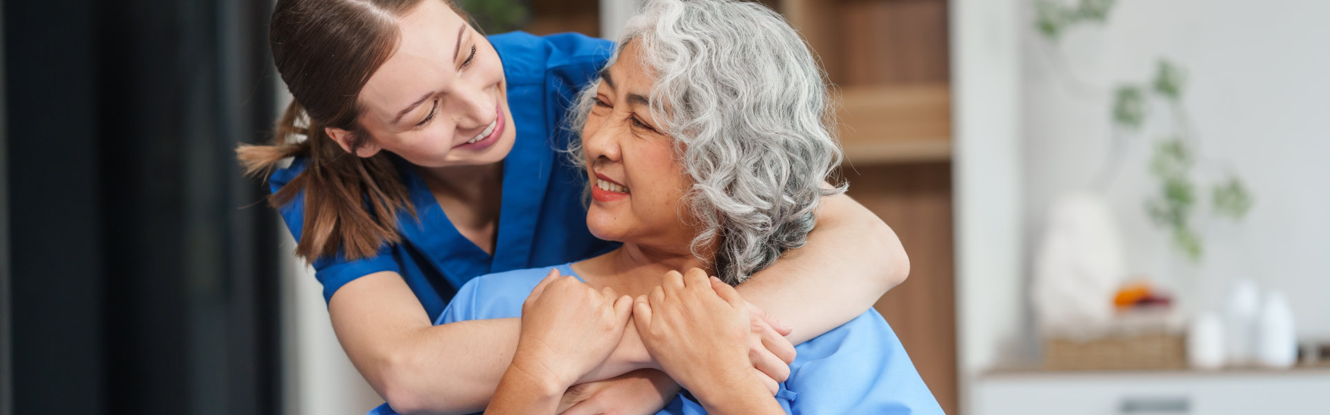 Young female caregiver embraces and smiles at elderly woman in wheelchair in a bright, homey indoor setting