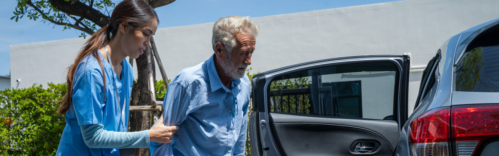 A healthcare worker in blue scrubs provides physical assistance to an elderly man with white hair as he exits a silver car