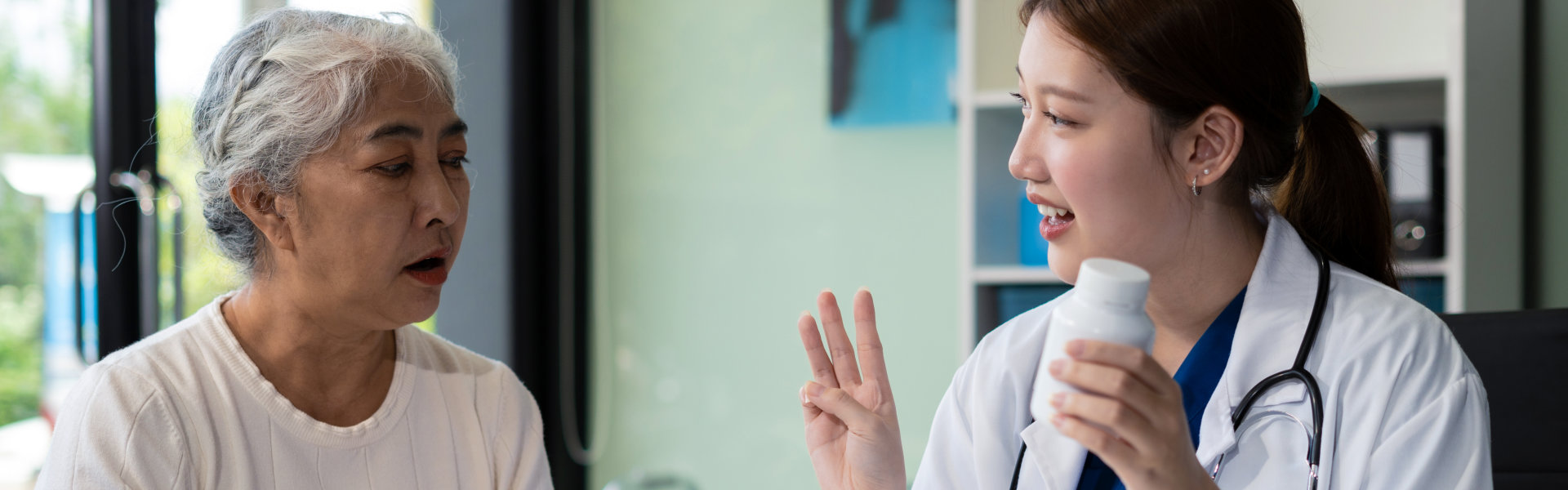 A doctor in a white coat and blue scrubs holds a bottle of medication while using her fingers to gesture the number three to an elderly patient during a consultation, with various medical supplies resting on the desk in front of them