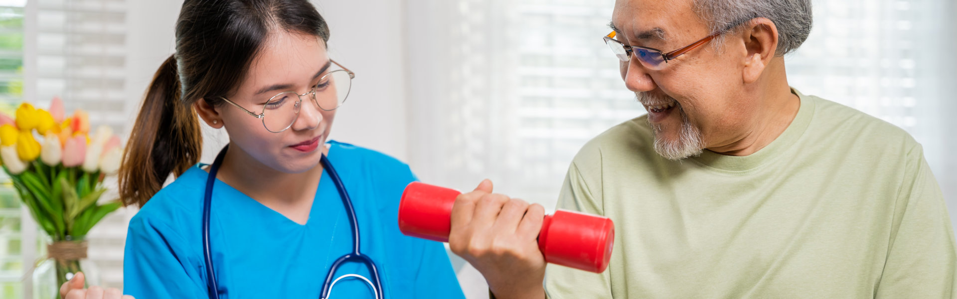 A caregiver in blue scrubs demonstrates an arm exercise while helping an elderly man who is smiling and lifting a red dumbbell