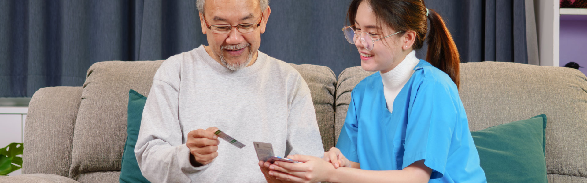 A friendly caregiver in a blue uniform sits on a sofa next to an elderly man, helping him review medical information or a card while medication and pharmacy boxes sit on the table in front of them