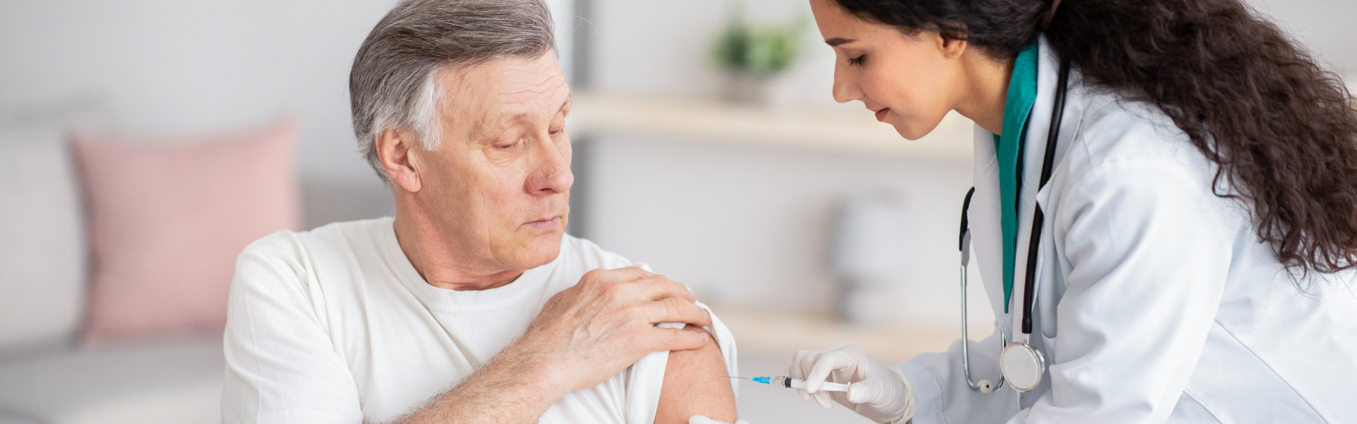 A doctor in a white coat and gloves administers an injection into the shoulder of an elderly man sitting in a chair